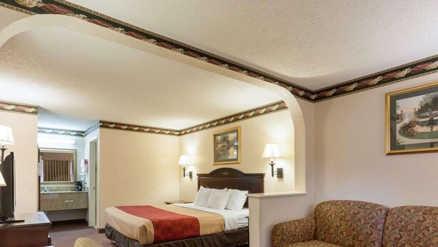 A hotel room with a bed, a sofa, a small table, a desk, a lamp, a painting on the wall, and a mirror. - Econo Lodge Grenada (Grenada)