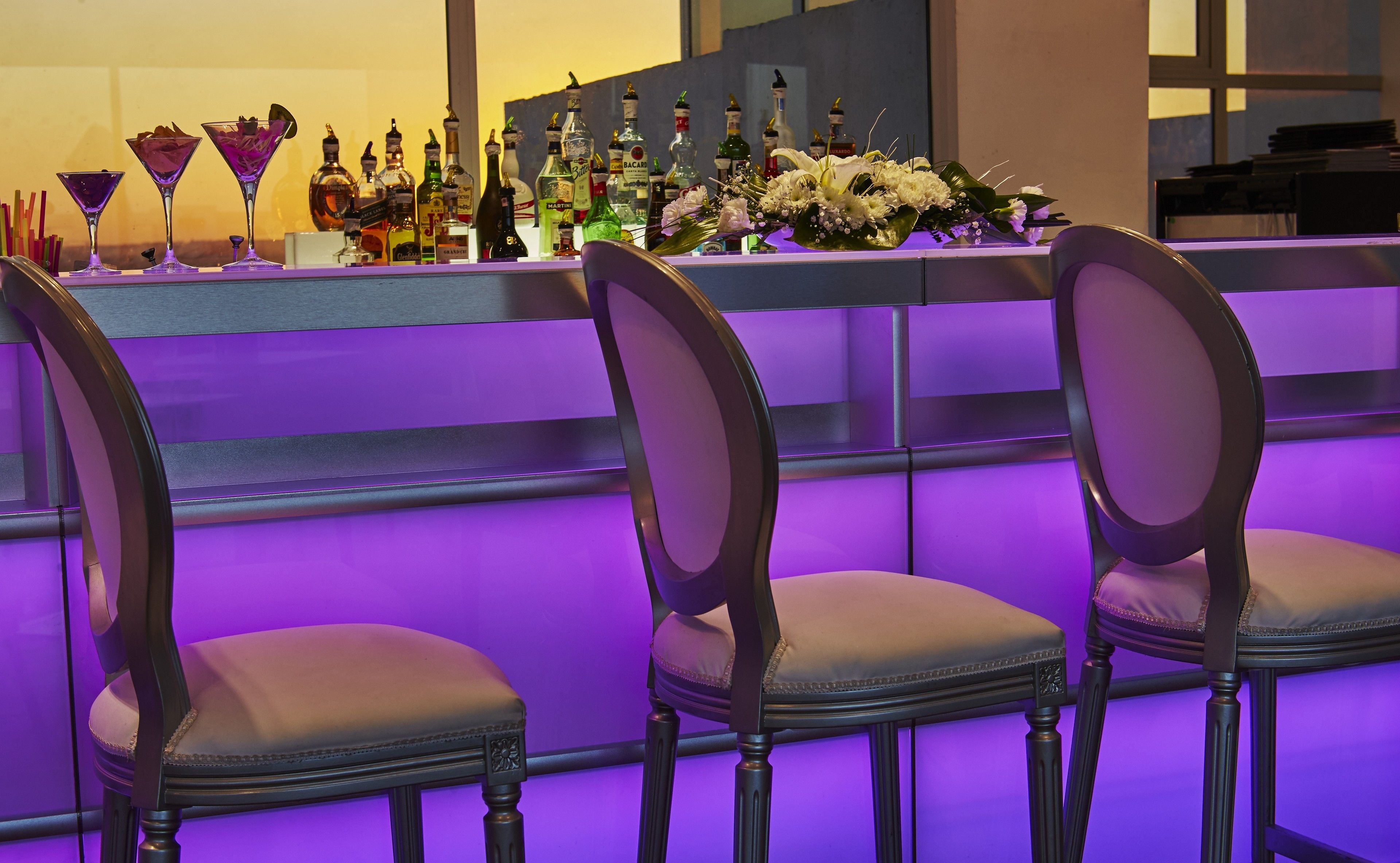 A bar area with a purple-lit counter, several chairs, and a variety of liquor bottles.