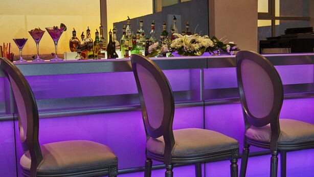 Zona de bar con mostrador iluminado en morado, varias sillas y una variedad de botellas de licor.