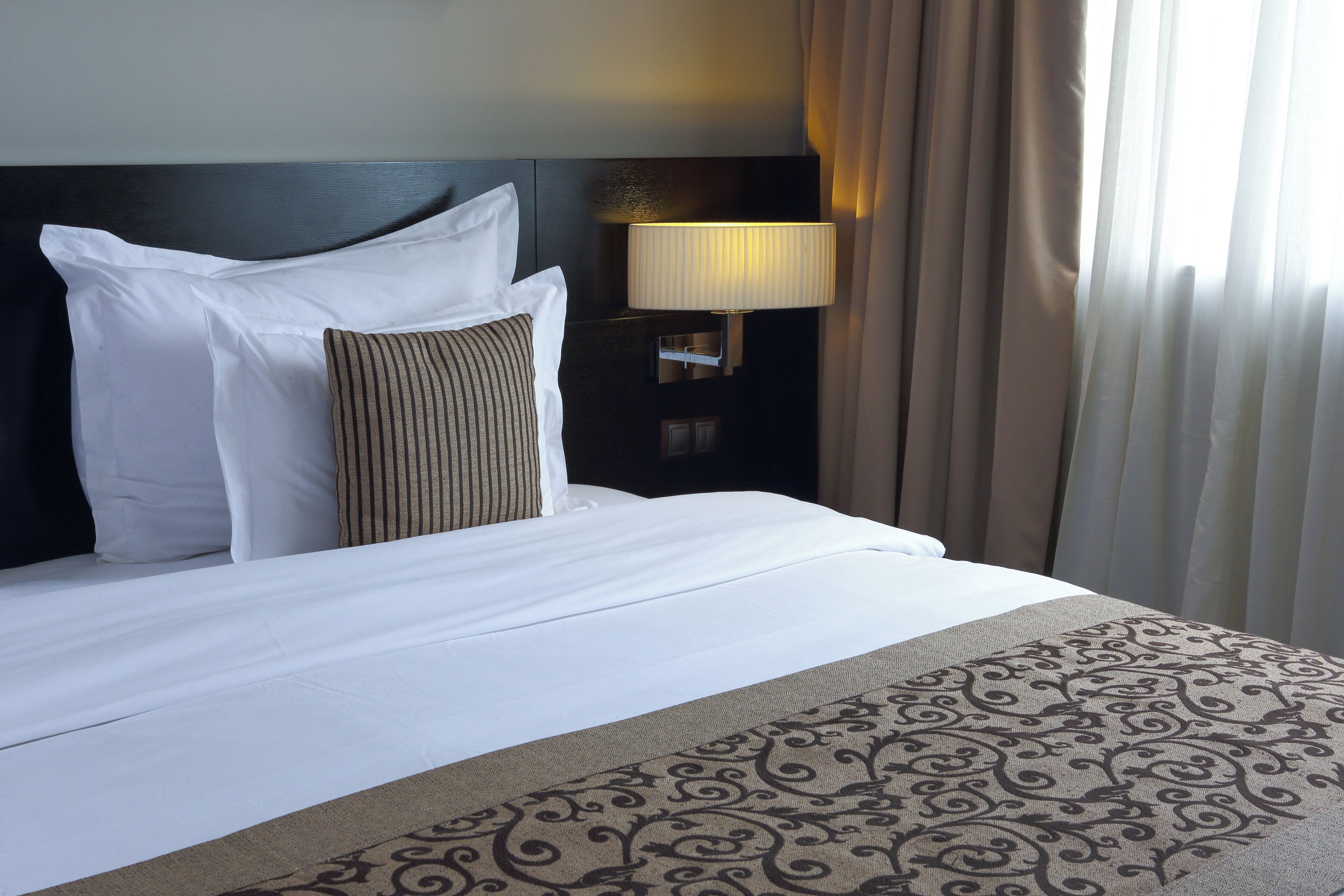A neatly made bed with white and striped pillows, a patterned throw, and a wall-mounted lamp.