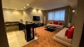 Una habitación de hotel moderna con sala de estar, un sofá con cojines rojos, una mesita de centro, un televisor de pantalla plana montado en la pared y una pequeña cocina con encimera de mármol.