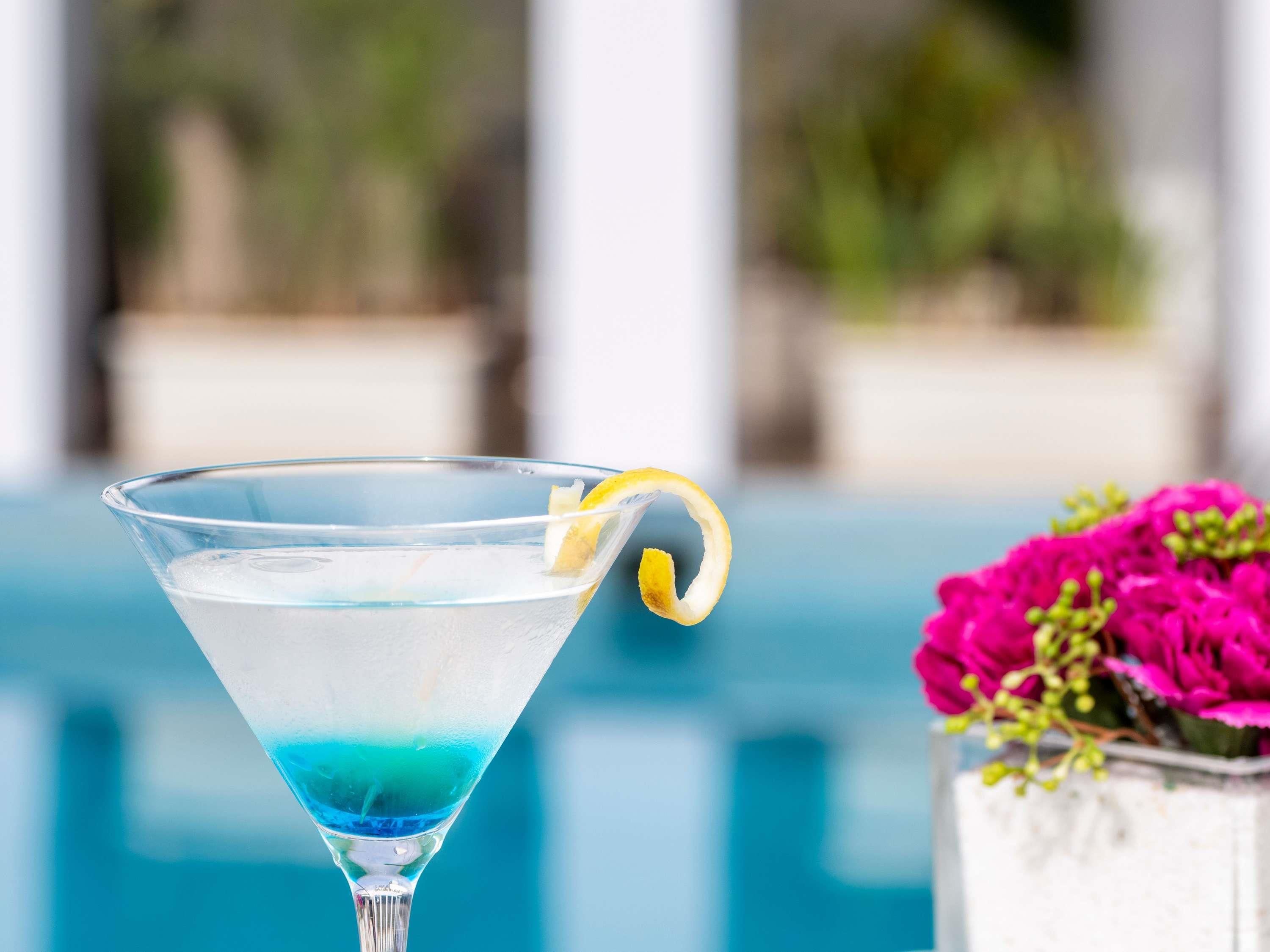 A clear glass martini glass with a lemon twist garnish, placed by a pool with a blurred background of greenery and a white structure.