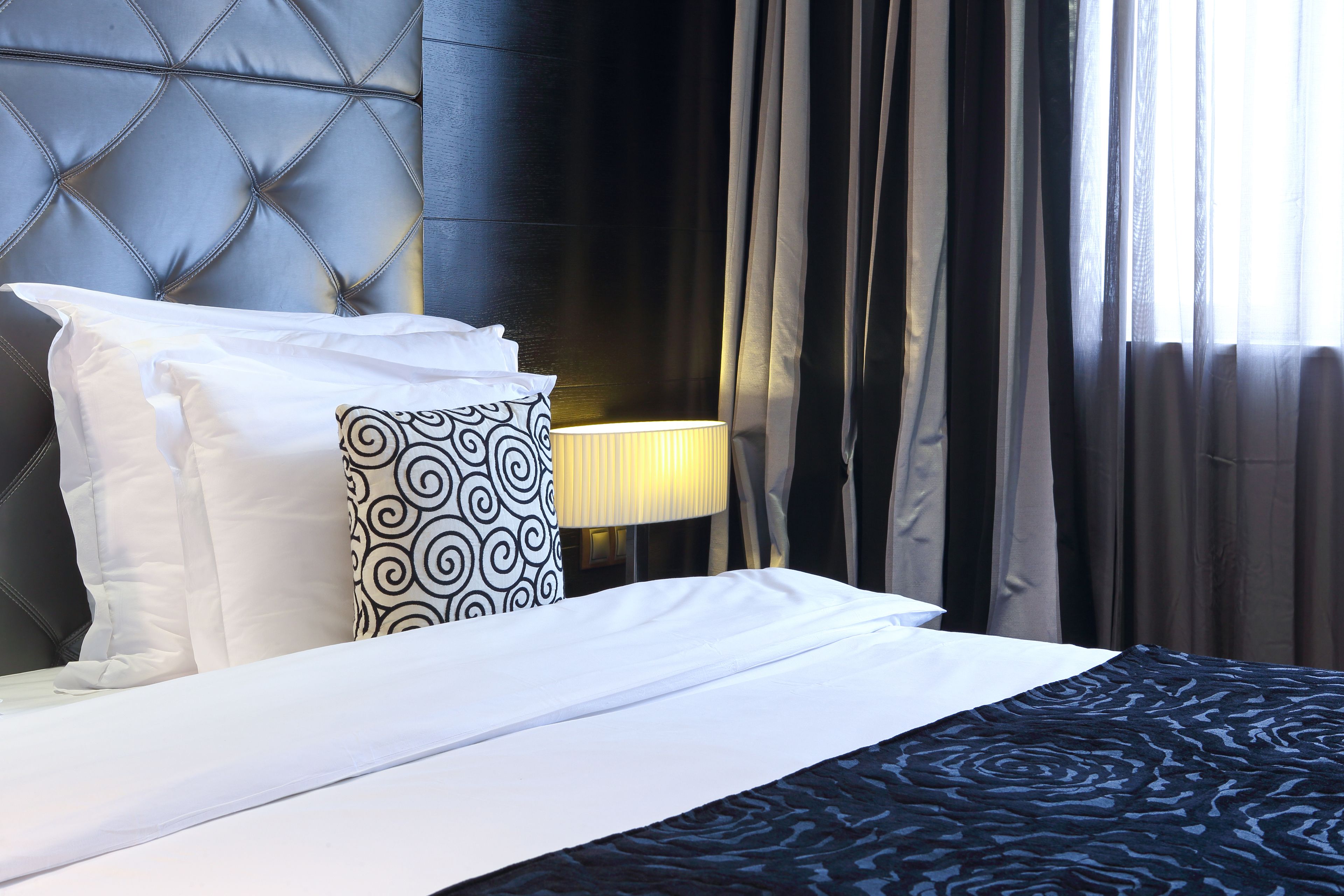 A hotel room with a tufted headboard, a bed with white and patterned pillows, a dark blue patterned bedspread, and a lamp on a bedside table.
