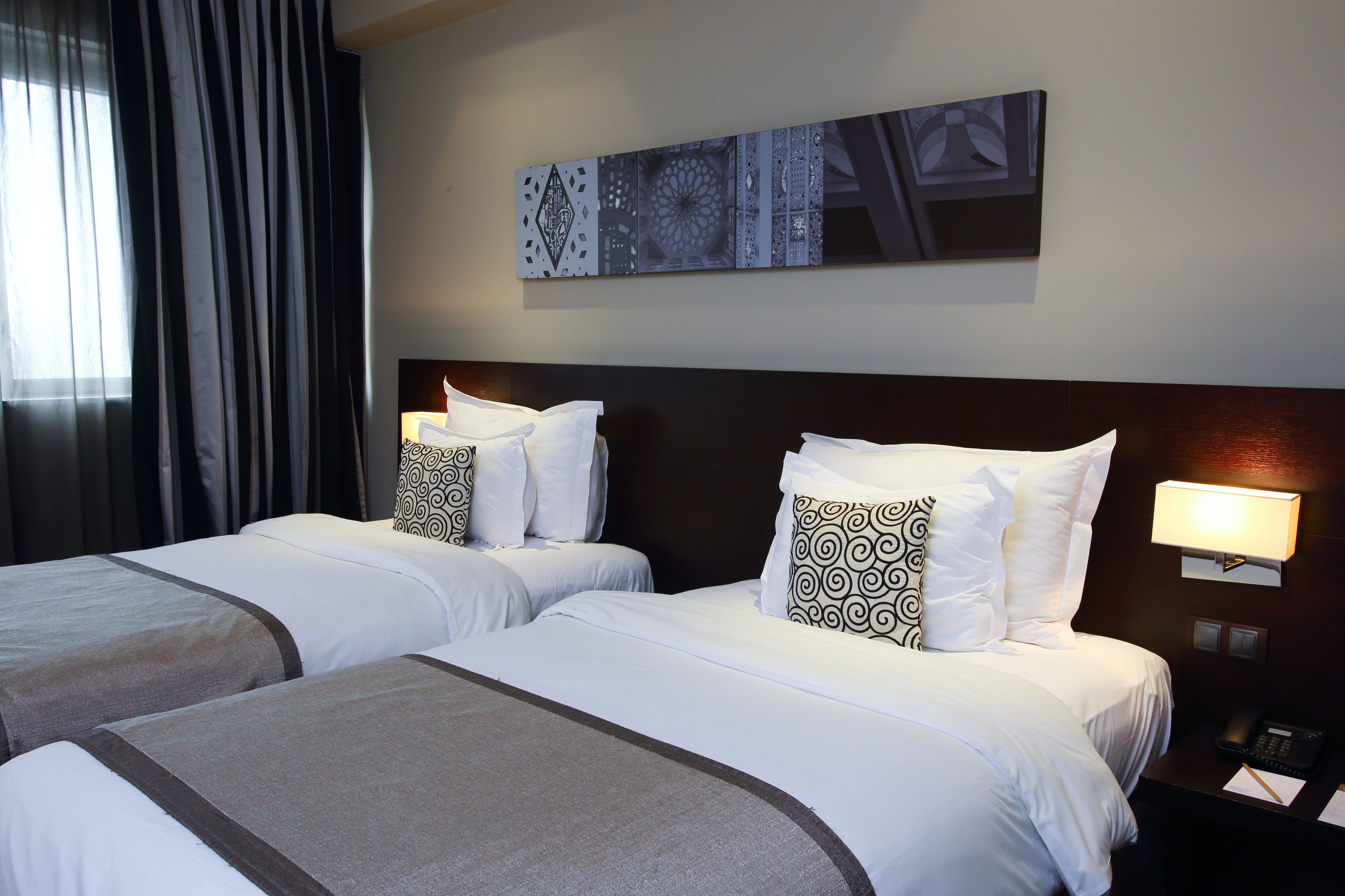 A hotel room with two beds, each with white bedding and patterned pillows, a dark headboard, a wall-mounted artwork, and a nightstand with a lamp and phone.