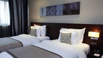 A hotel room with two beds, each with white bedding and patterned pillows, a dark headboard, a wall-mounted artwork, and a nightstand with a lamp and phone.