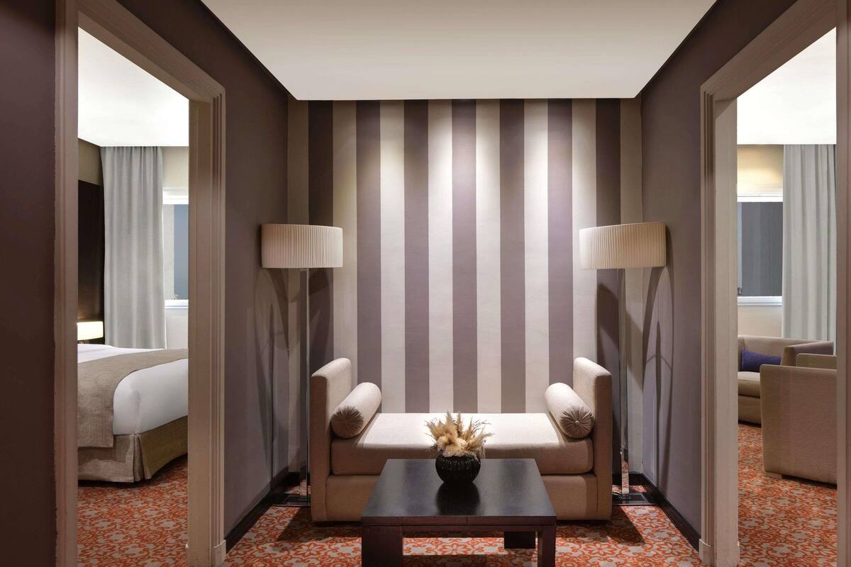 A hotel room with a striped wall, a sofa, two armchairs, a coffee table, and a bed.
