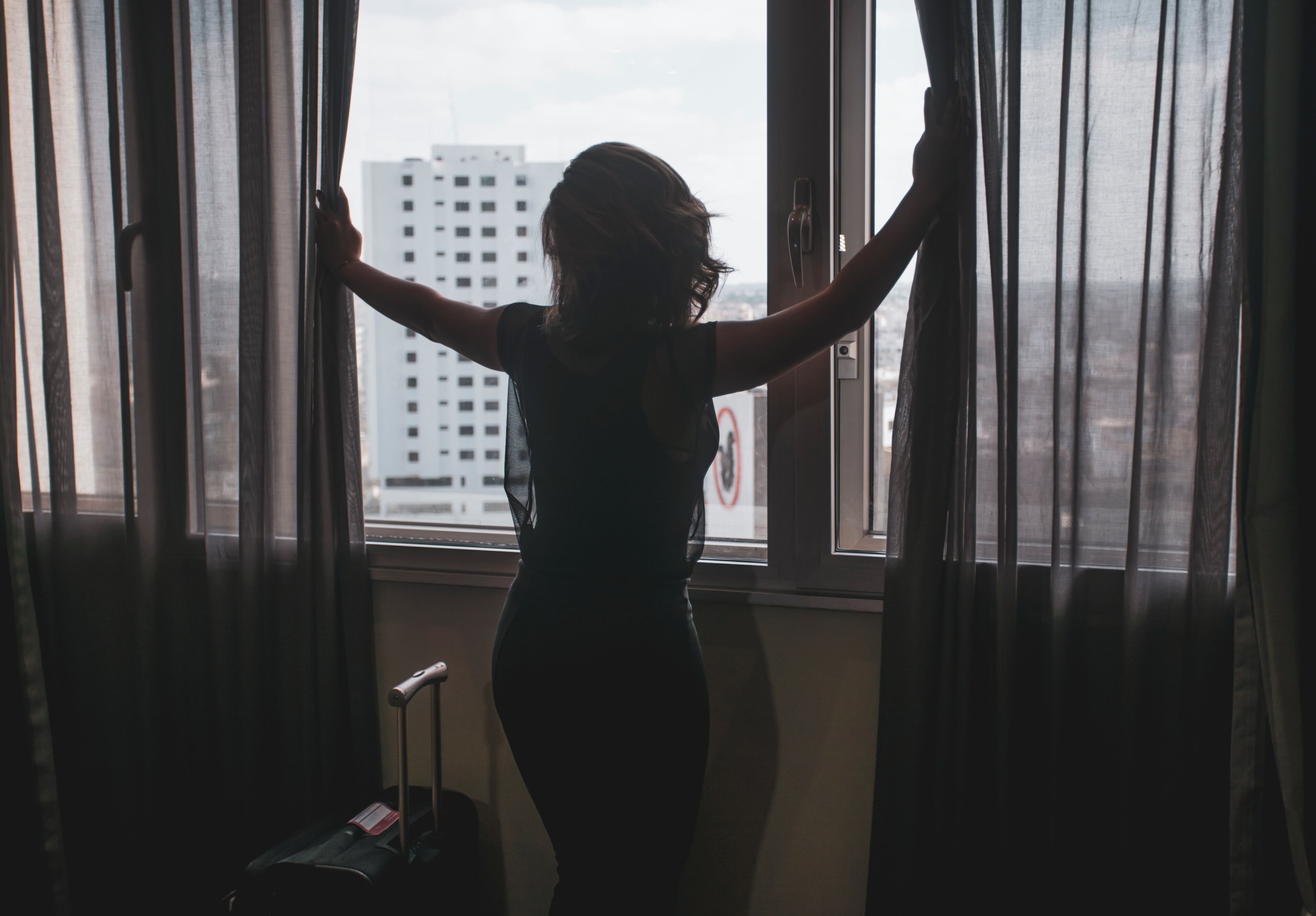 A person standing by a window with curtains, a suitcase, and a door handle visible.