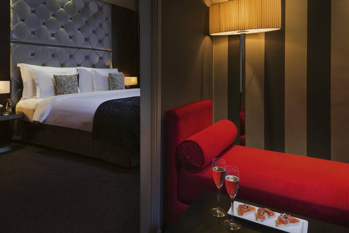 A hotel room with a tufted headboard bed, a red sofa, a table with two glasses of champagne, and a plate of food.
