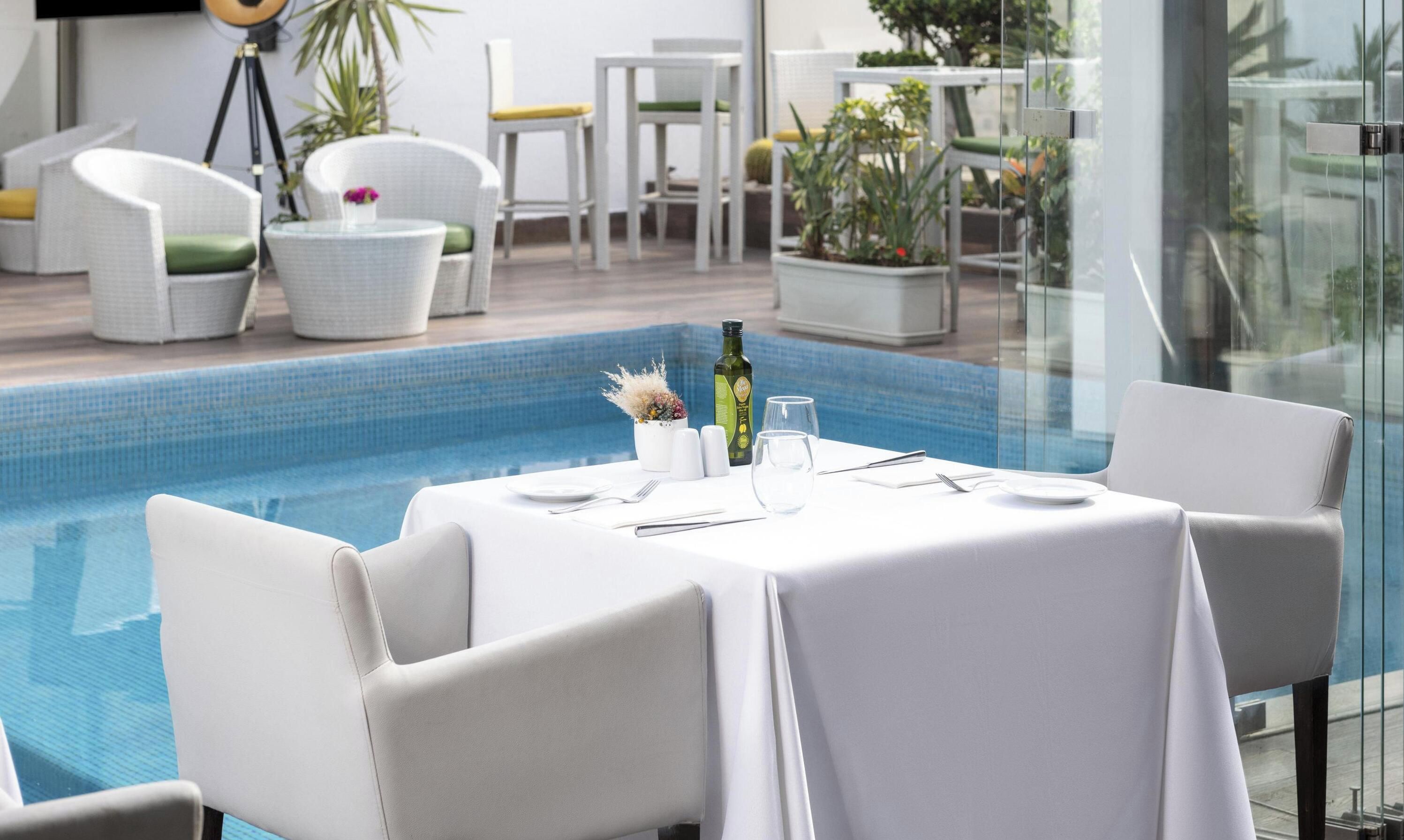 Un espace repas aménagé au bord de la piscine, avec une table dressée pour deux personnes, entourée de chaises blanches, une bouteille d’huile d’olive, des salières et des poivrières, et un verre.