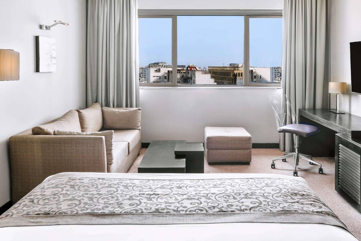 A hotel room with a bed, a sofa, a desk, a chair, and a view of the cityscape.