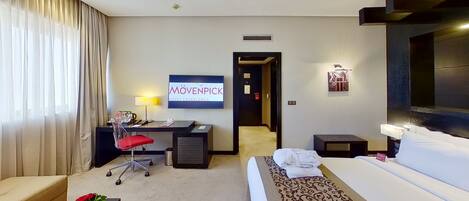 Habitación de hotel con cama, escritorio, silla y televisión mostrando el nombre del hotel.