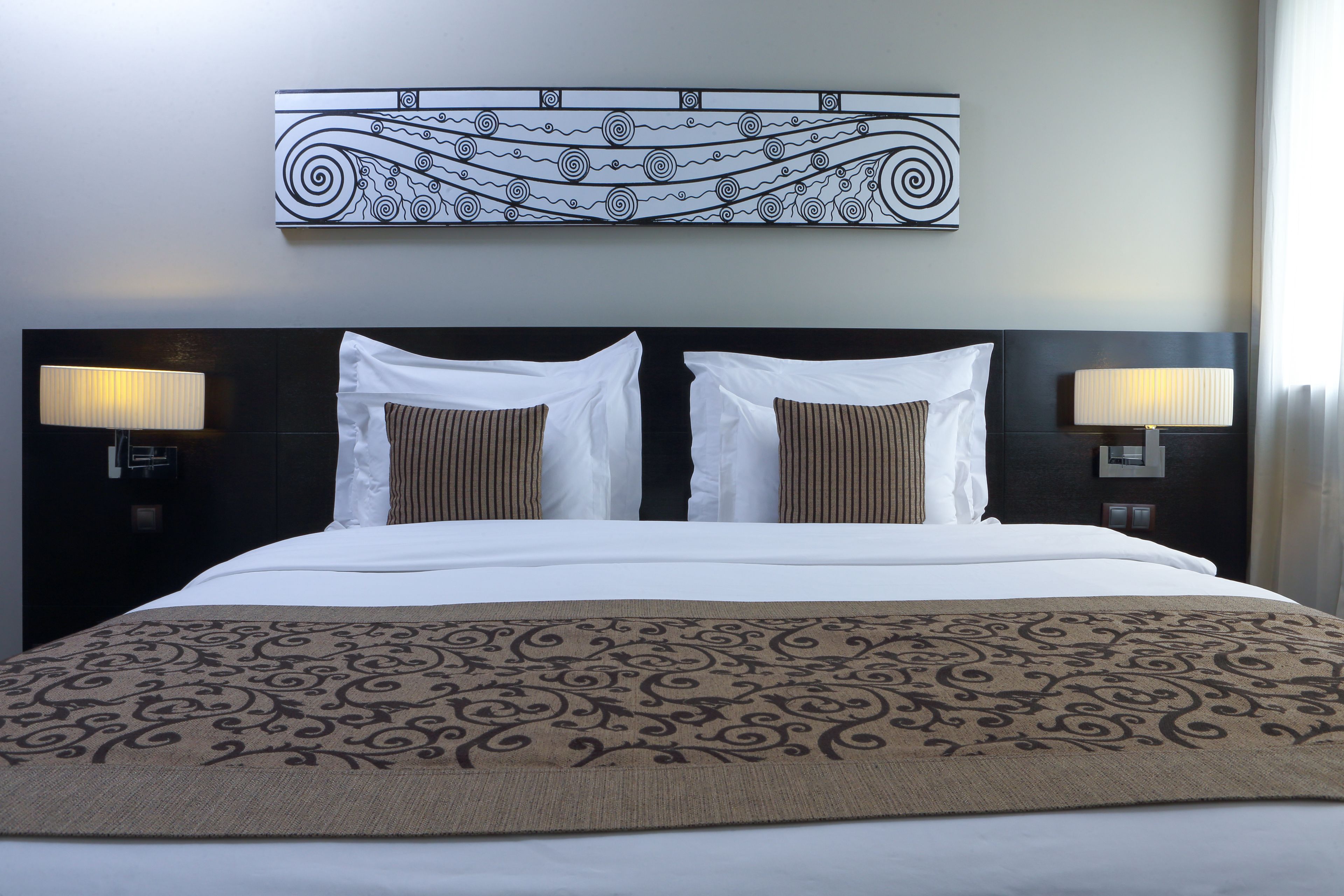 A hotel room with a large bed, two bedside lamps, a decorative wall piece, and a patterned throw blanket.