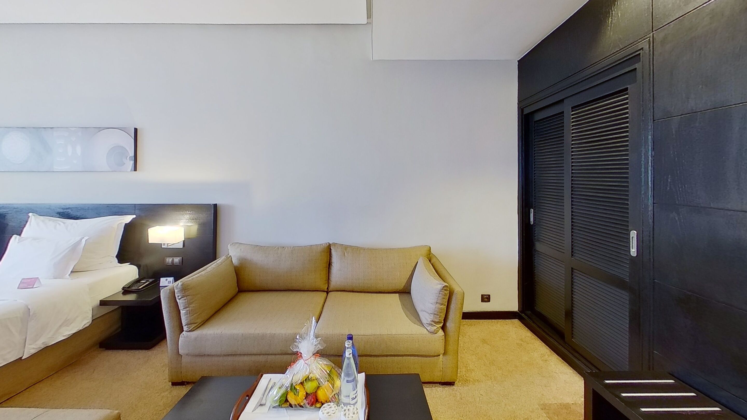 A hotel room with a bed, a sofa, a coffee table with fruits and a bottle, and a dark wardrobe.