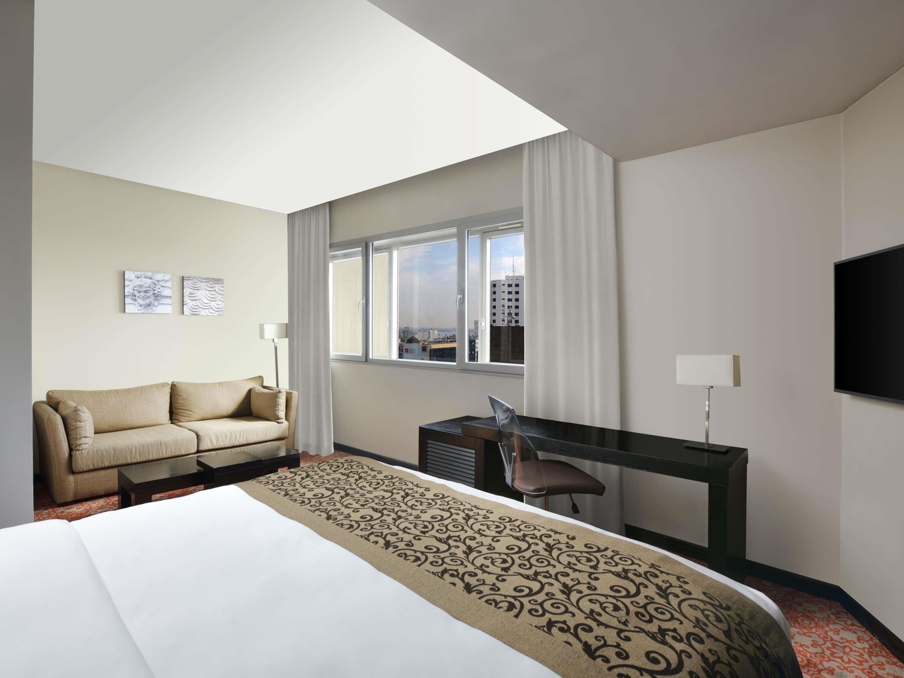 A hotel room with a bed, a desk, a chair, a sofa, and a TV.