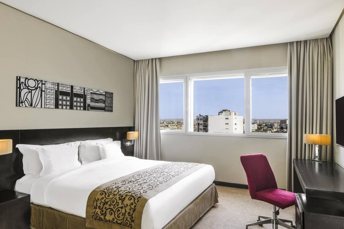 A hotel room with a large bed, a desk, a chair, and a view of the city through the window.