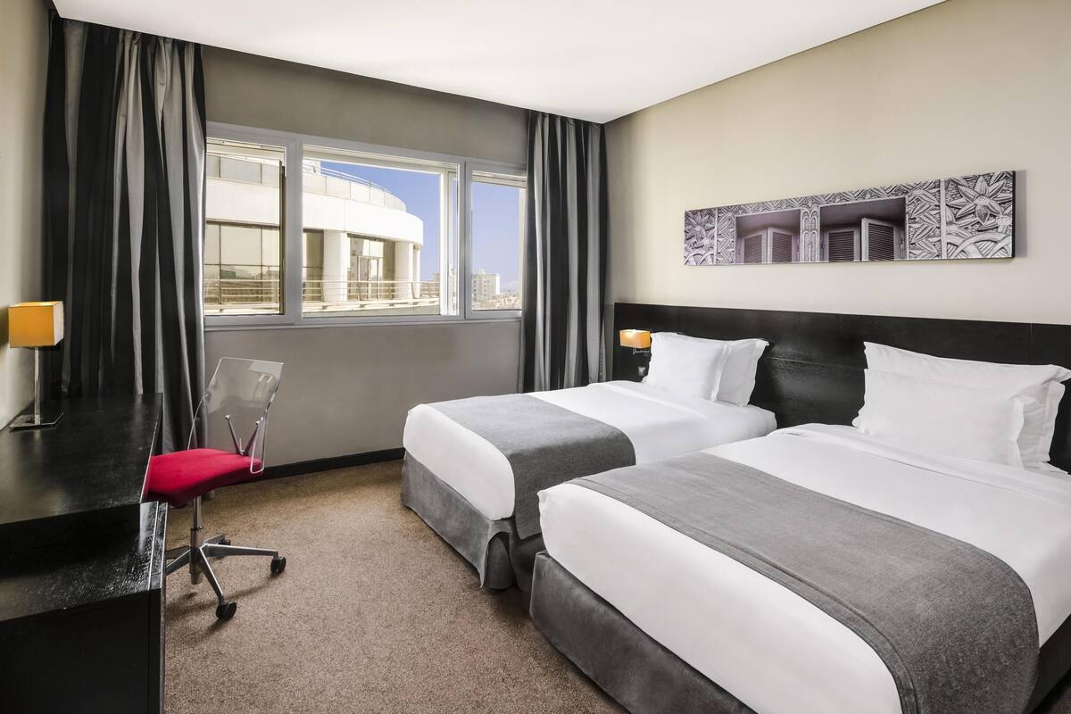 A hotel room with two beds, a desk, a chair, and a window with a view of buildings.