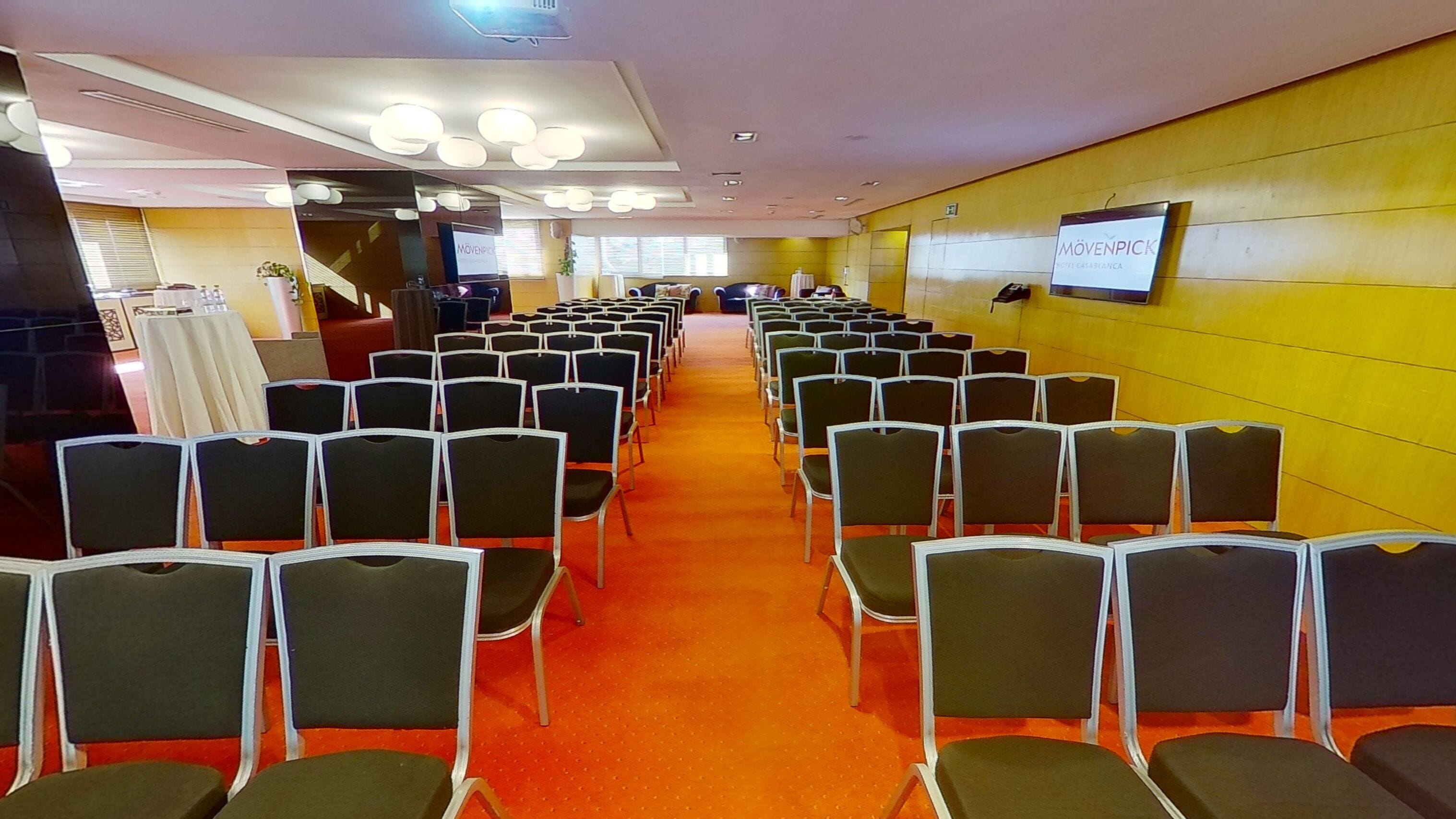 Un salón de conferencias con filas de sillas, piso alfombrado y una pared amarilla con una pantalla que muestra texto.