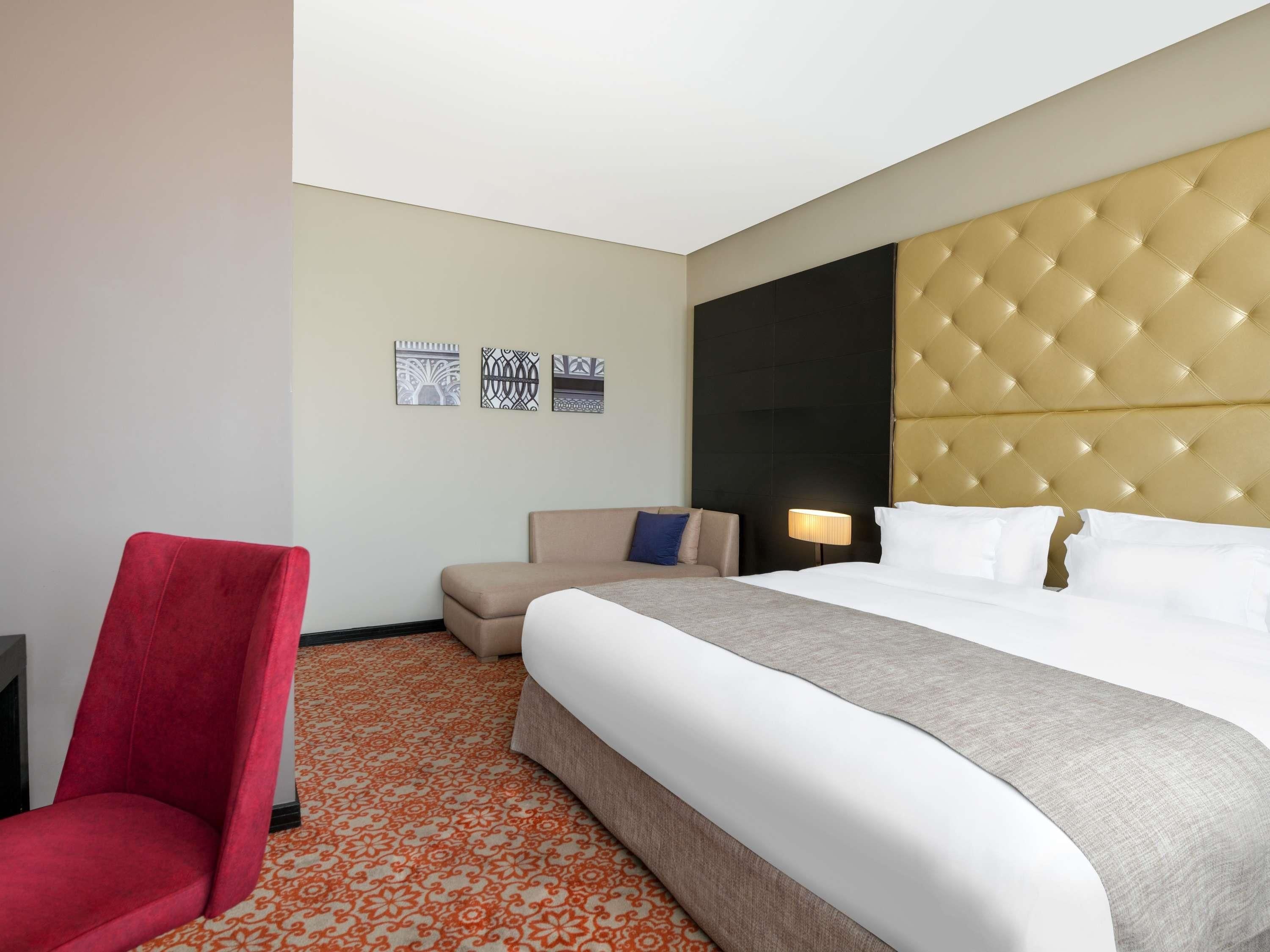 A hotel room with a bed, a red chair, a small sofa, and patterned carpet.