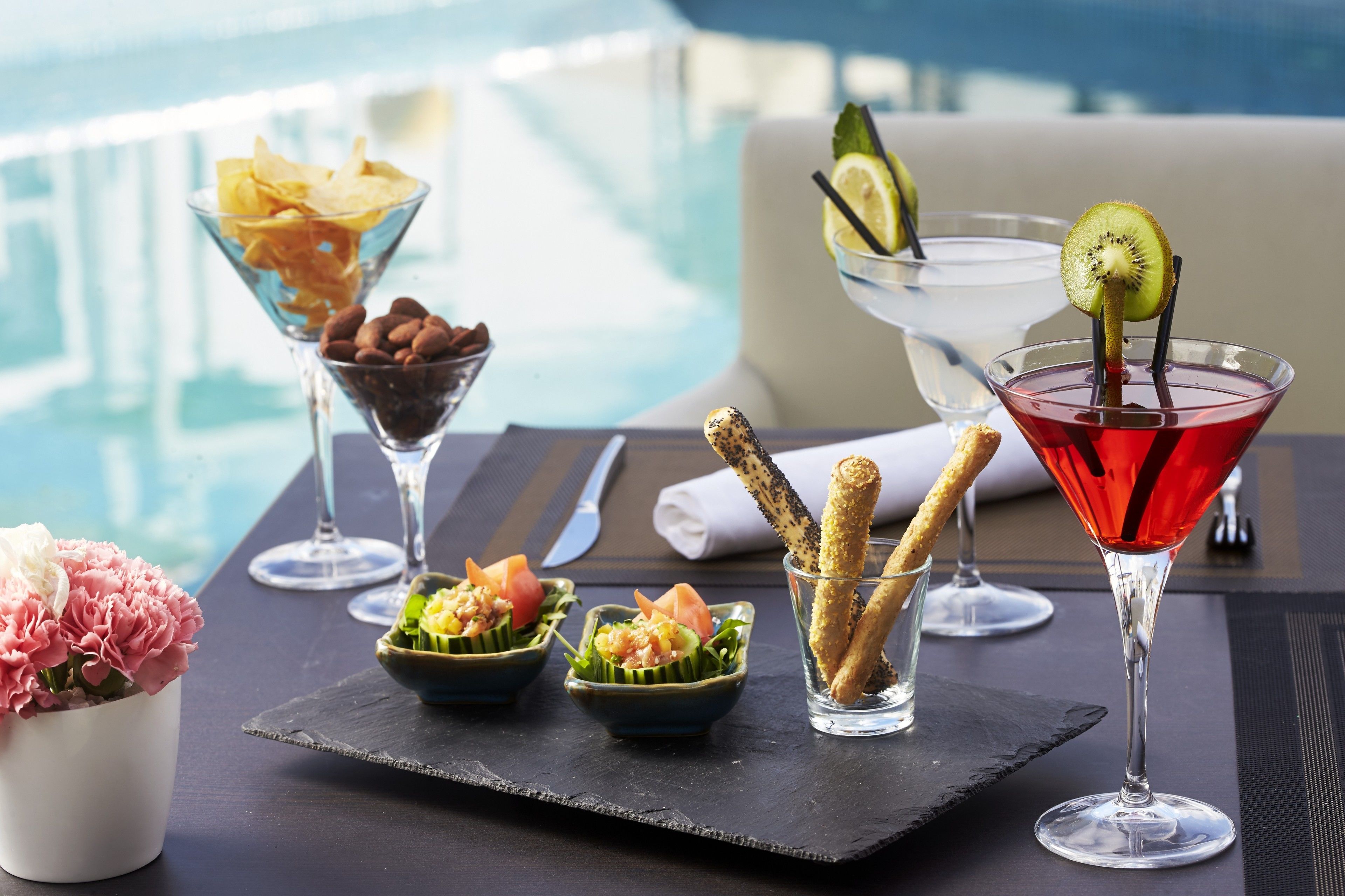 A table with various dishes and drinks, including a red cocktail with a kiwi slice, a bowl of chips, and a small dish with a salad.