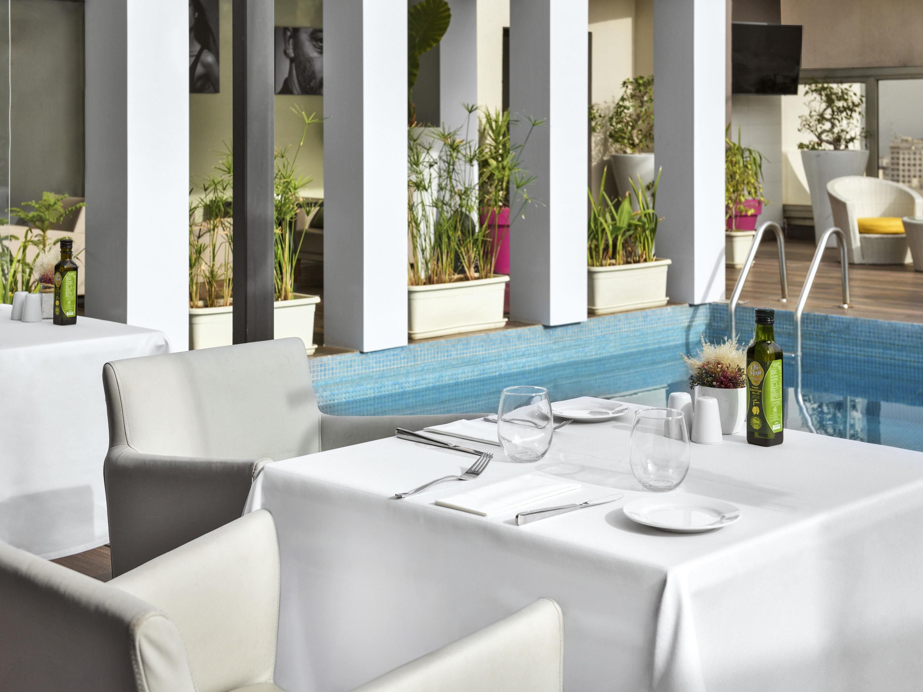 Comedor con mesas dispuestas para dos, plantas en macetas y una piscina al fondo.