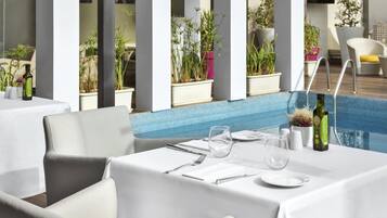 Comedor con mesas dispuestas para dos, plantas en macetas y una piscina al fondo.