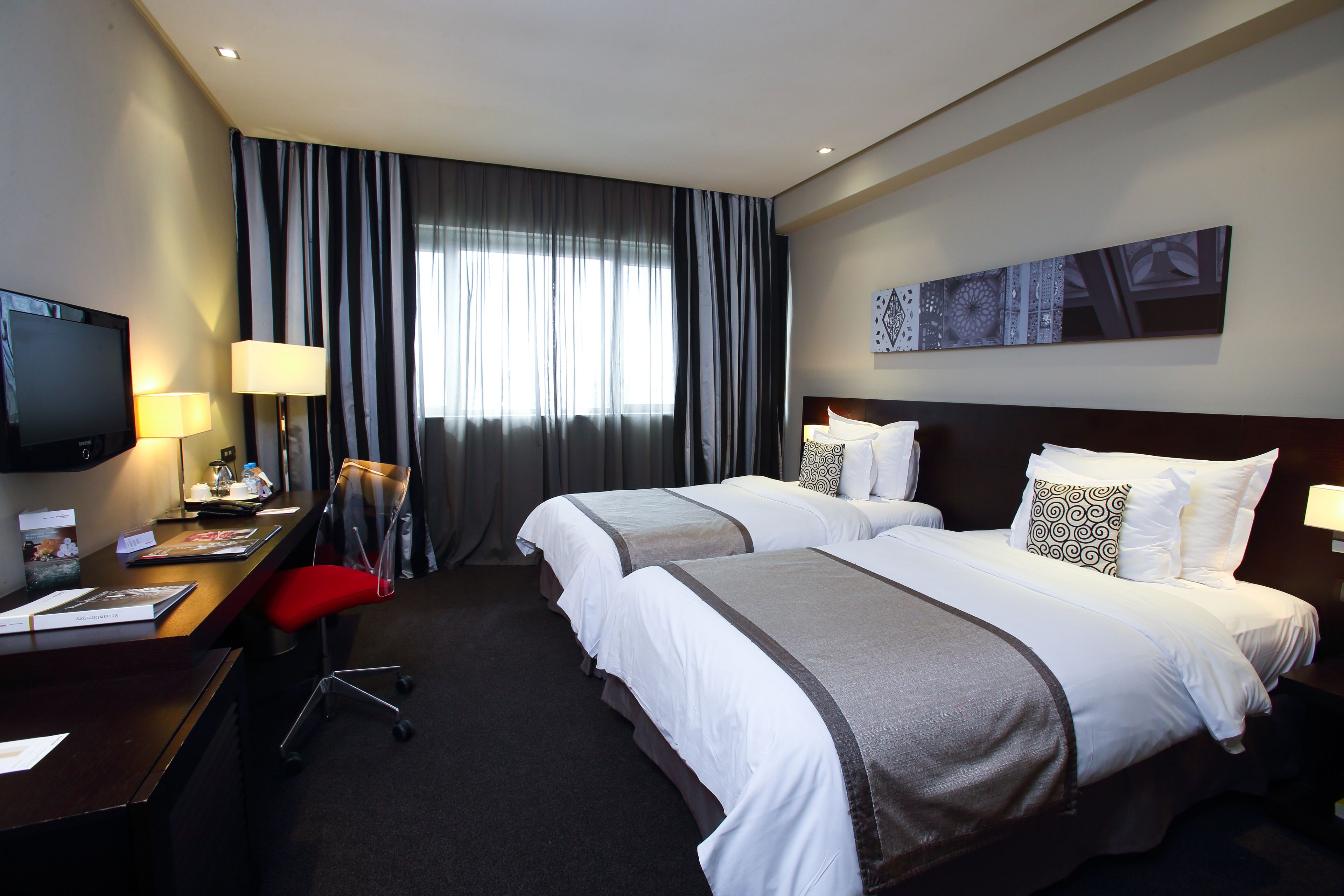 Habitación de hotel con dos camas, un escritorio con computadora y lámpara, televisor y un ventanal con cortinas.