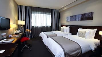 Habitación de hotel con dos camas, un escritorio con computadora y lámpara, televisor y un ventanal con cortinas.