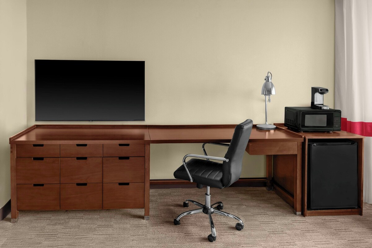 Een houten bureau met lades, een zwarte bureaustoel, een televisie, een lamp en een kleine koelkast.