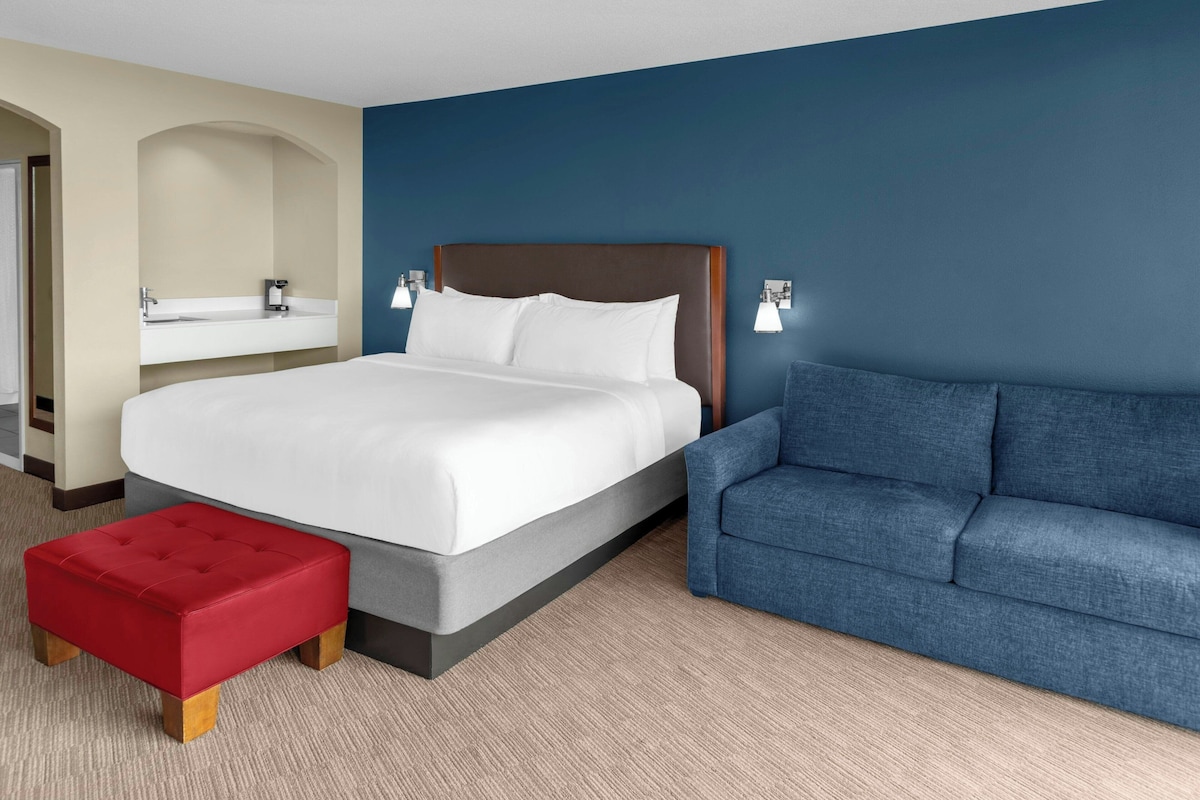 A hotel room with a bed, a red ottoman, a blue sofa, a bathroom with a sink and mirror, and a blue accent wall.
