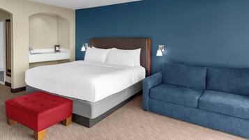 A hotel room with a bed, a red ottoman, a blue sofa, a bathroom with a sink and mirror, and a blue accent wall.