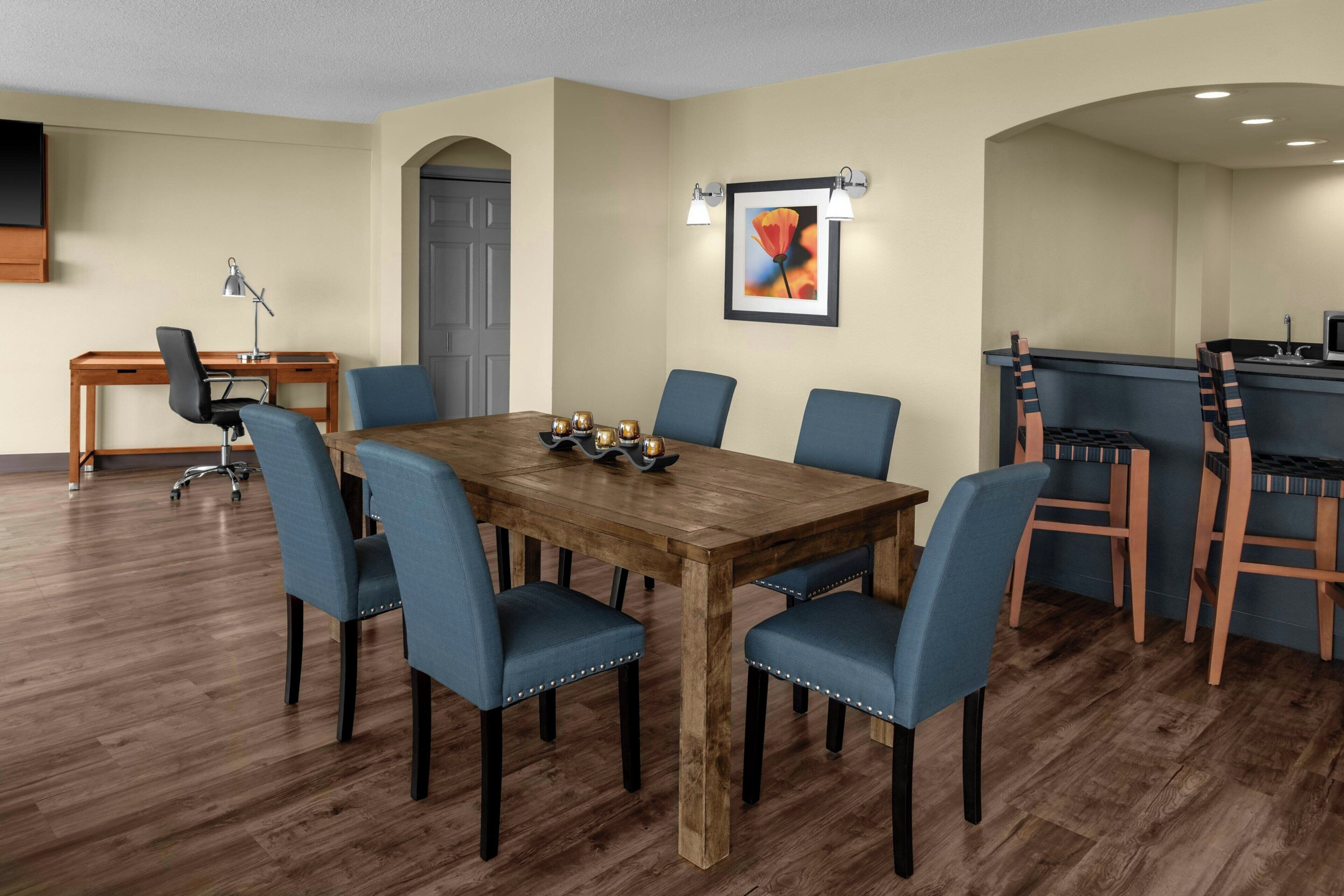 A dining area with a wooden table, blue chairs, a desk with a lamp, and a framed picture on the wall.
