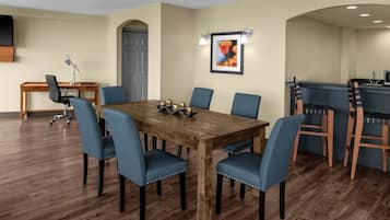 A dining area with a wooden table, blue chairs, a desk with a lamp, and a framed picture on the wall.