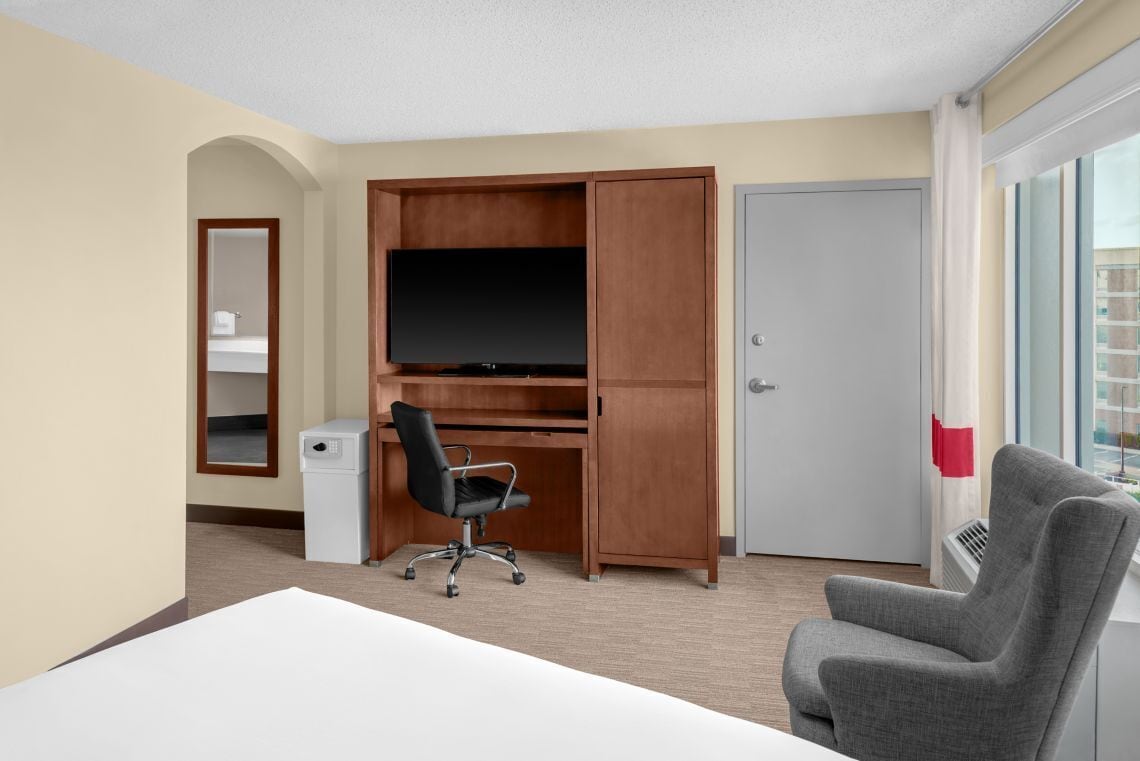 Quarto de hotel com cama, escrivaninha com televisão, cadeira, guarda-roupa e porta.