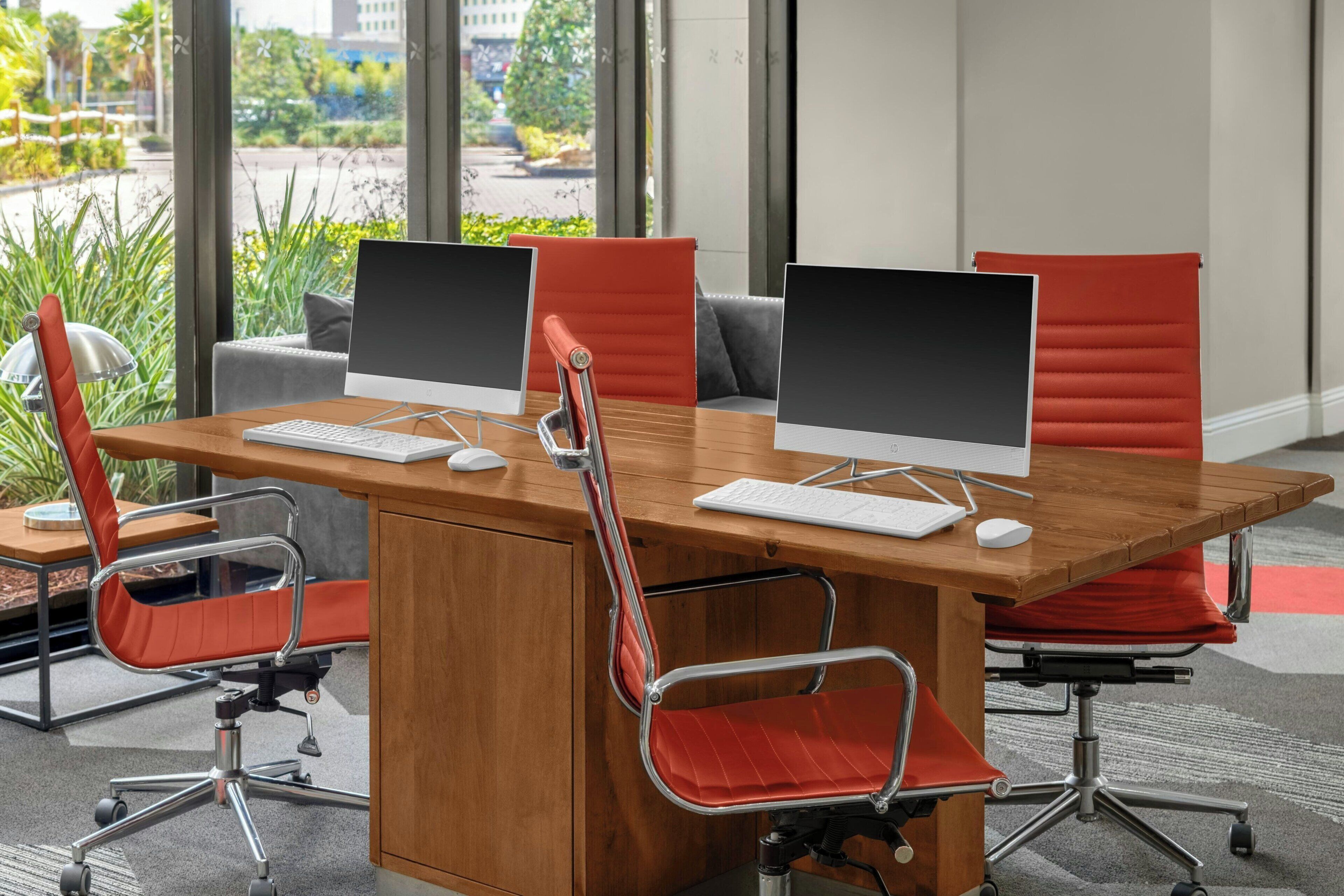 A conference room with two desks, each with a computer monitor, keyboard, and mouse. There are two red office chairs with wheels.
