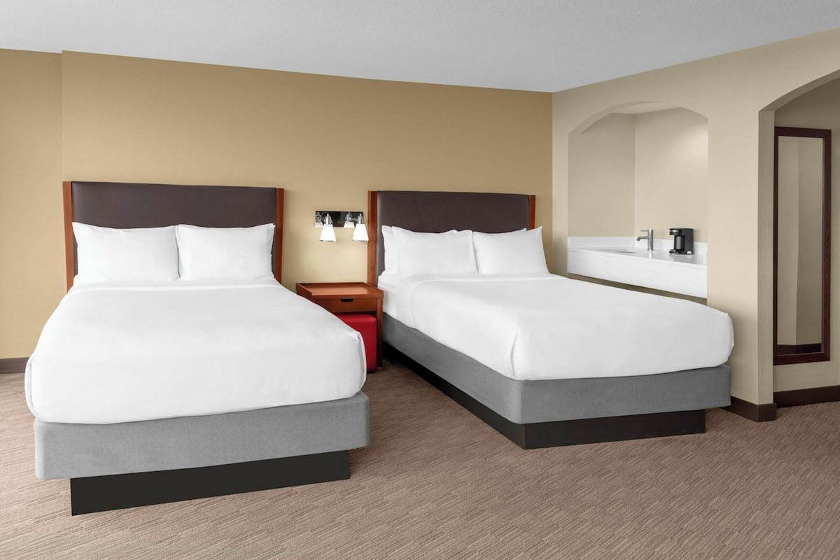 A hotel room with two beds, a nightstand, a red suitcase, and a bathroom with a mirror and sink.