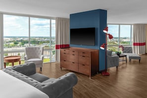 Una habitación de hotel moderna con un ventanal grande, un televisor de pantalla plana, un aparador de madera y una zona de estar con sofá y sillones.