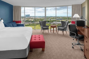 Habitación de hotel con una cama grande, dos sillones, un escritorio con computadora y vista a la ciudad.