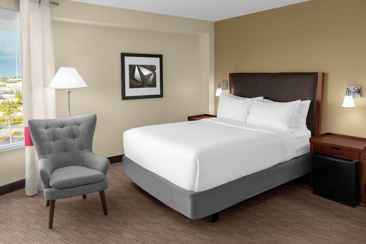 Een hotelkamer met een bed, een stoel, een nachtkastje, een lamp en een ingelijst kunstwerk aan de muur.