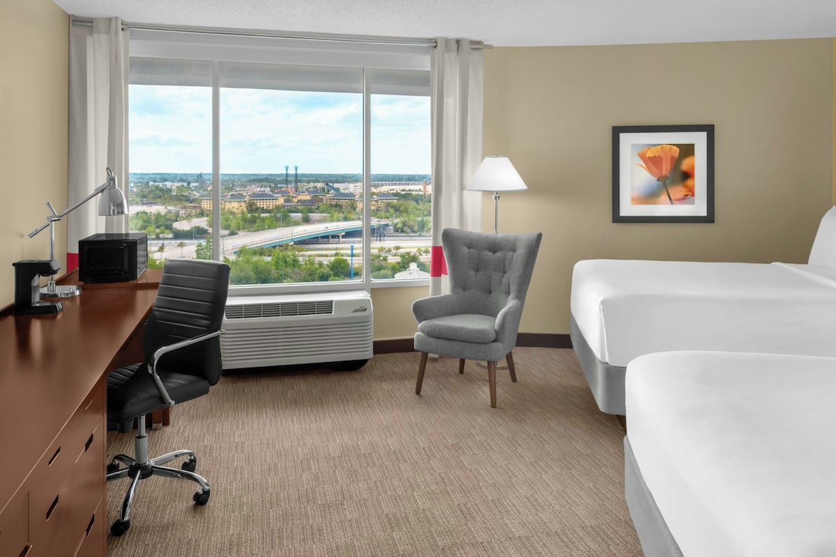 Een hotelkamer met een groot raam, een bureau met stoel, een bed en een lijst met een afbeelding aan de muur.