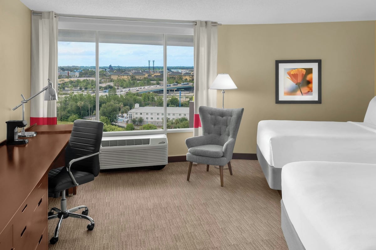 Een hotelkamer met een bed, een bureau met een stoel, een staande lamp en een groot raam met uitzicht op de stad.