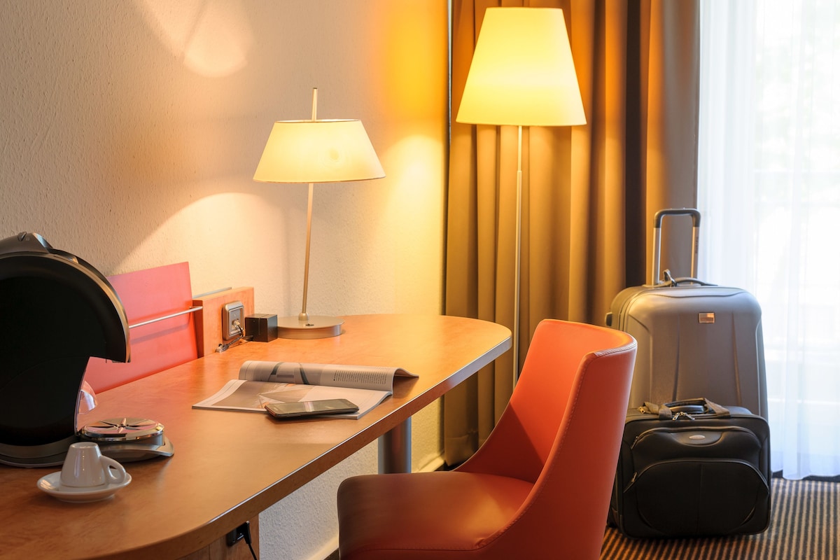 Una habitación de hotel con escritorio, lámpara, silla, maleta y cama.