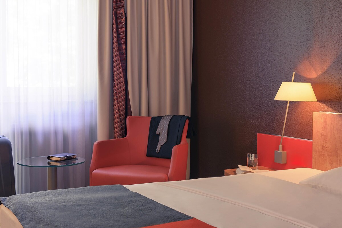 A hotel room with a bed, a red armchair, a small side table, a lamp, and a view of the outside through curtains.