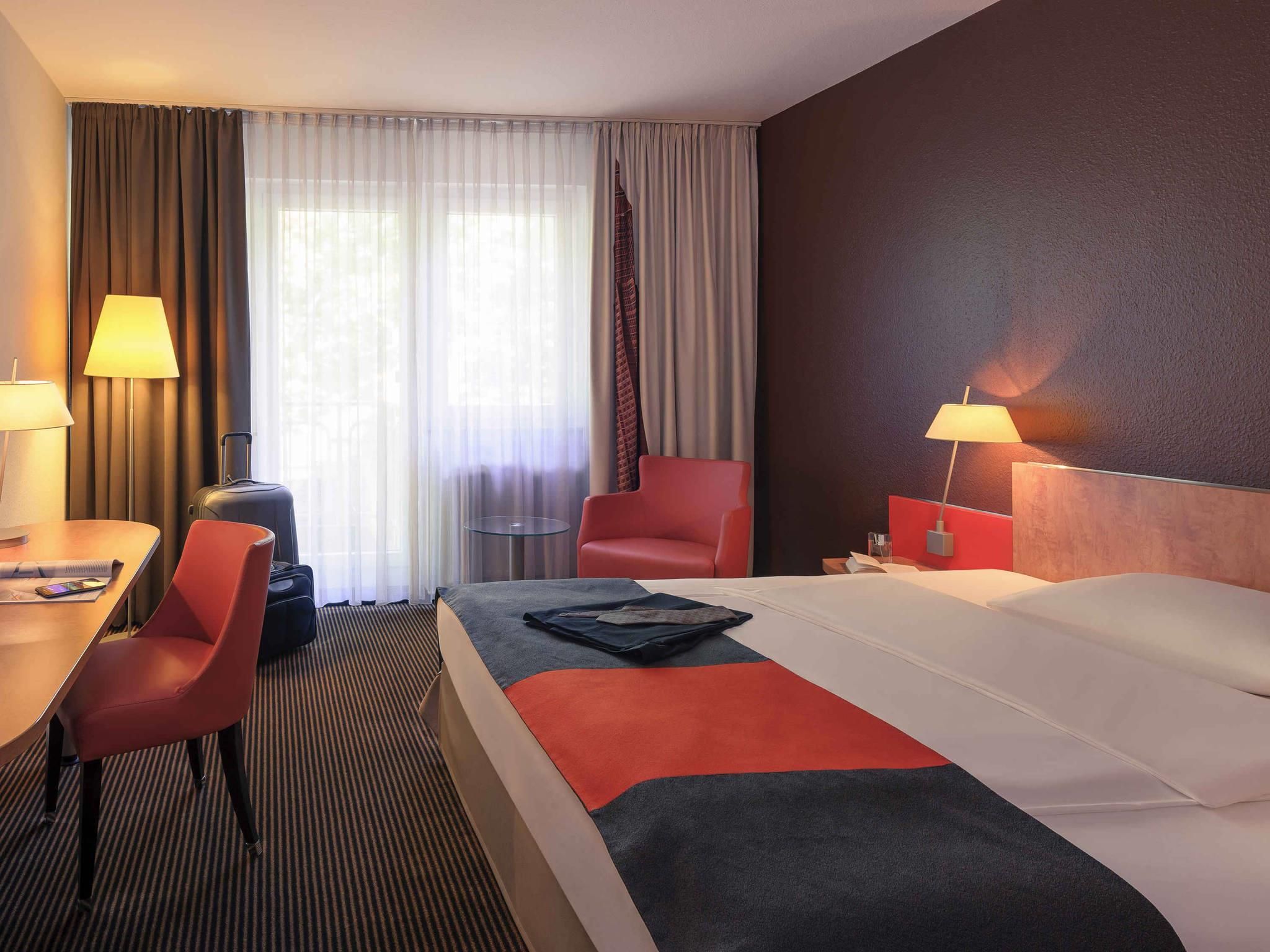 Une chambre d’hôtel avec un lit, un bureau, une chaise, une lampe et une fenêtre avec des rideaux.
