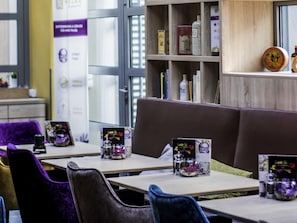 Interior de restaurante con mesas de madera, sillas tapizadas en morado y negro, y una estantería con diversos objetos.