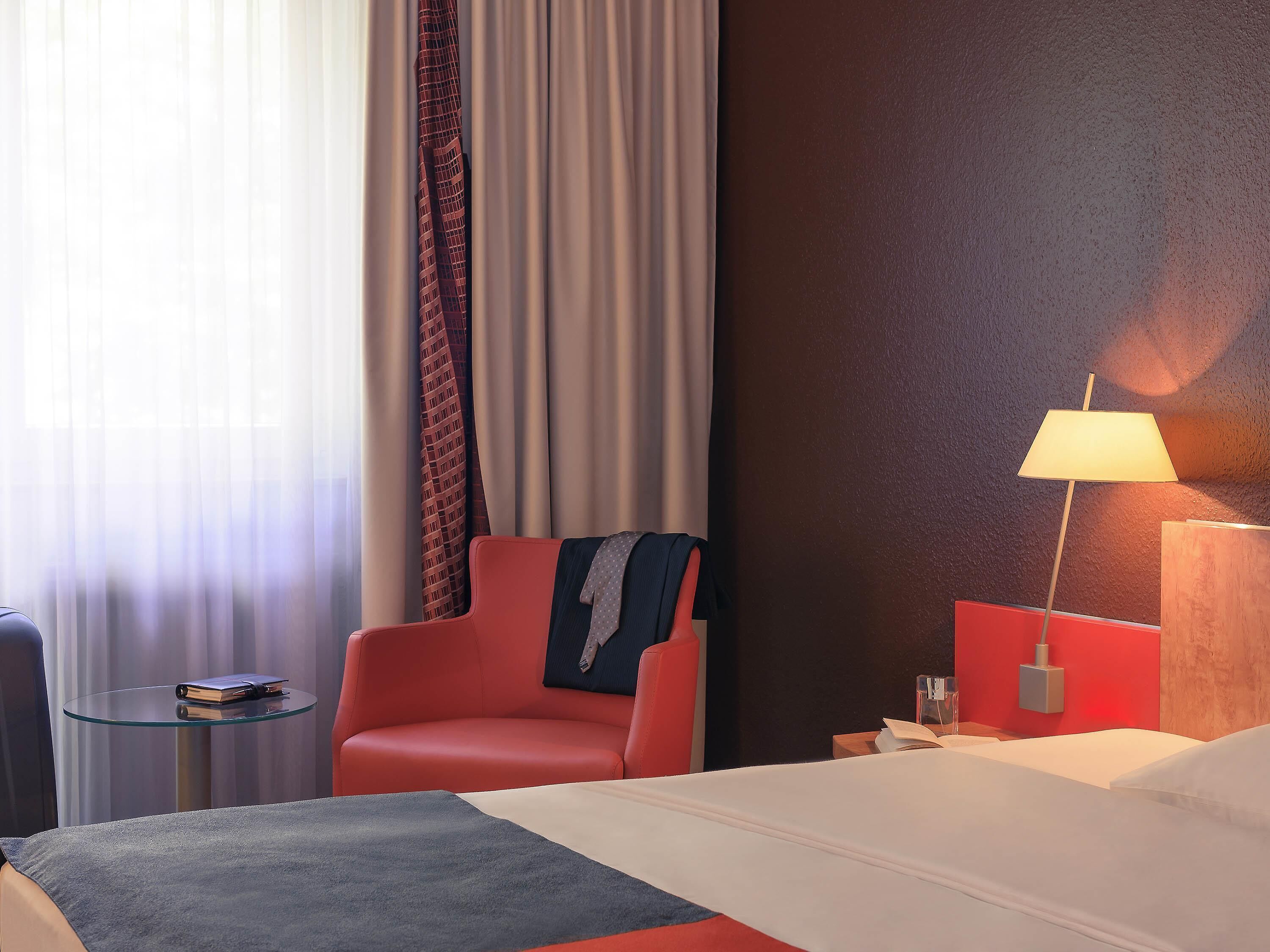 Ein Hotelzimmer mit einem Bett, einem roten Sessel, einem kleinen Nachttisch, einer Lampe und Blick durch Vorhänge nach draußen.