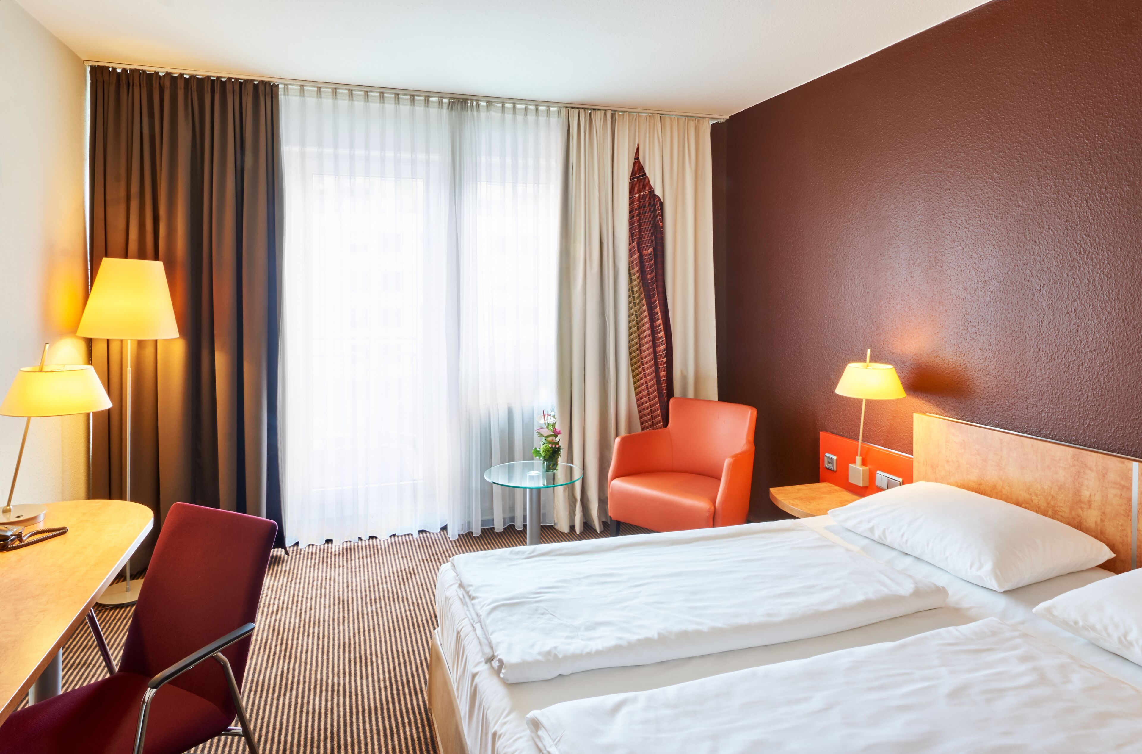 Habitación de hotel con dos camas, un escritorio, una silla, una mesa con lámpara y vista a un edificio por la ventana.