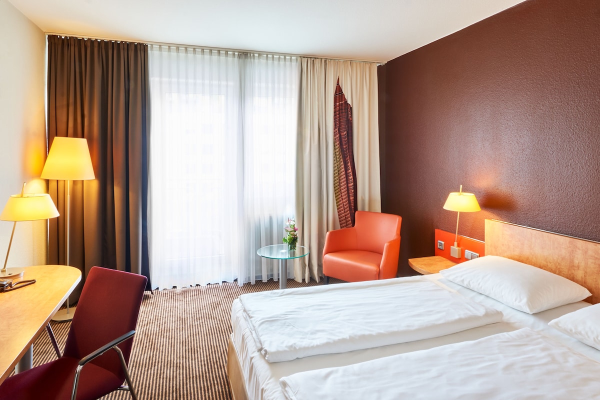 Habitación de hotel con dos camas, un escritorio, una silla, una mesa con lámpara y vista a un edificio por la ventana.