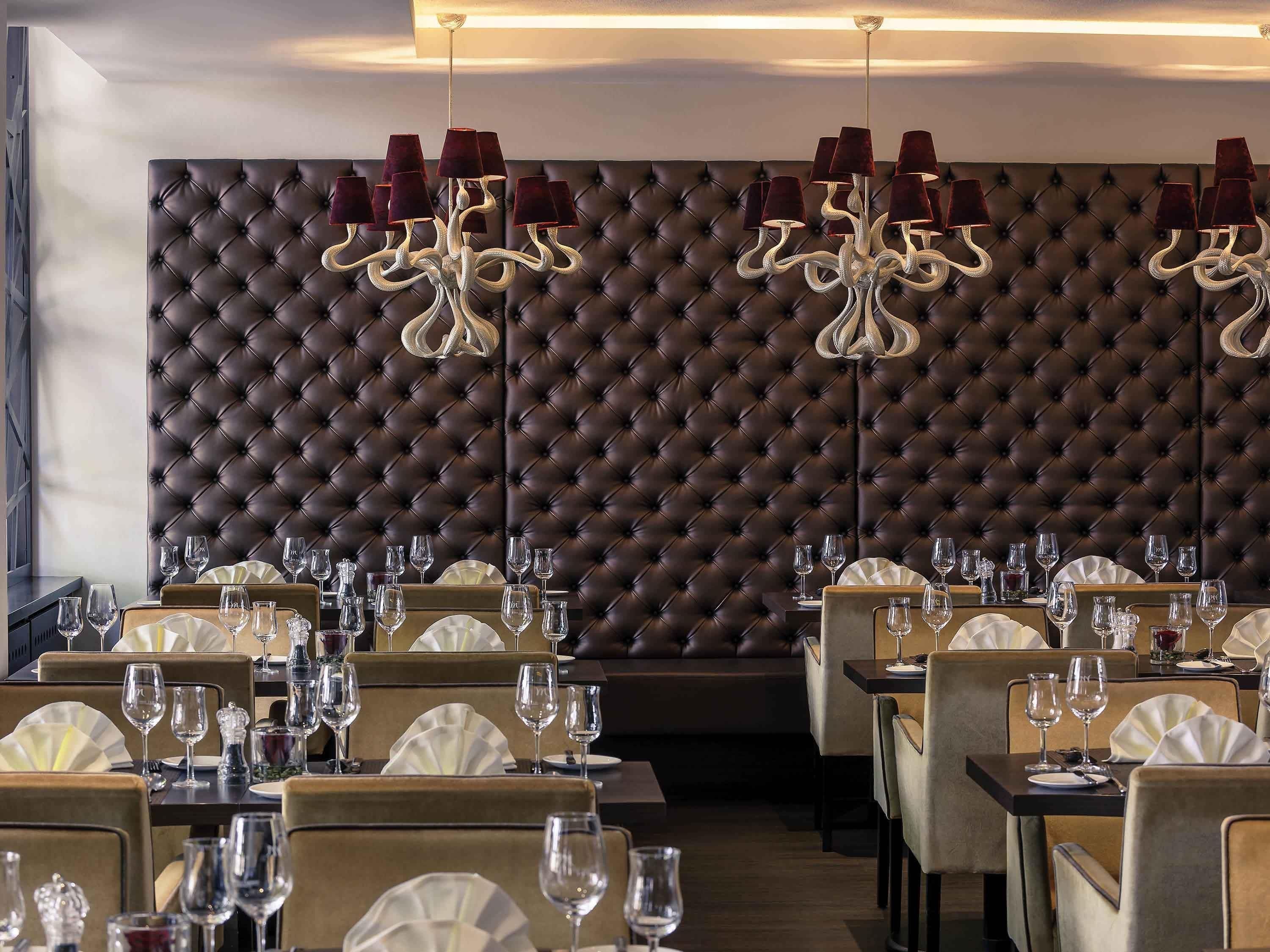 A restaurant with a tufted leather wall, multiple tables set with wine glasses, and a chandelier with red shades.