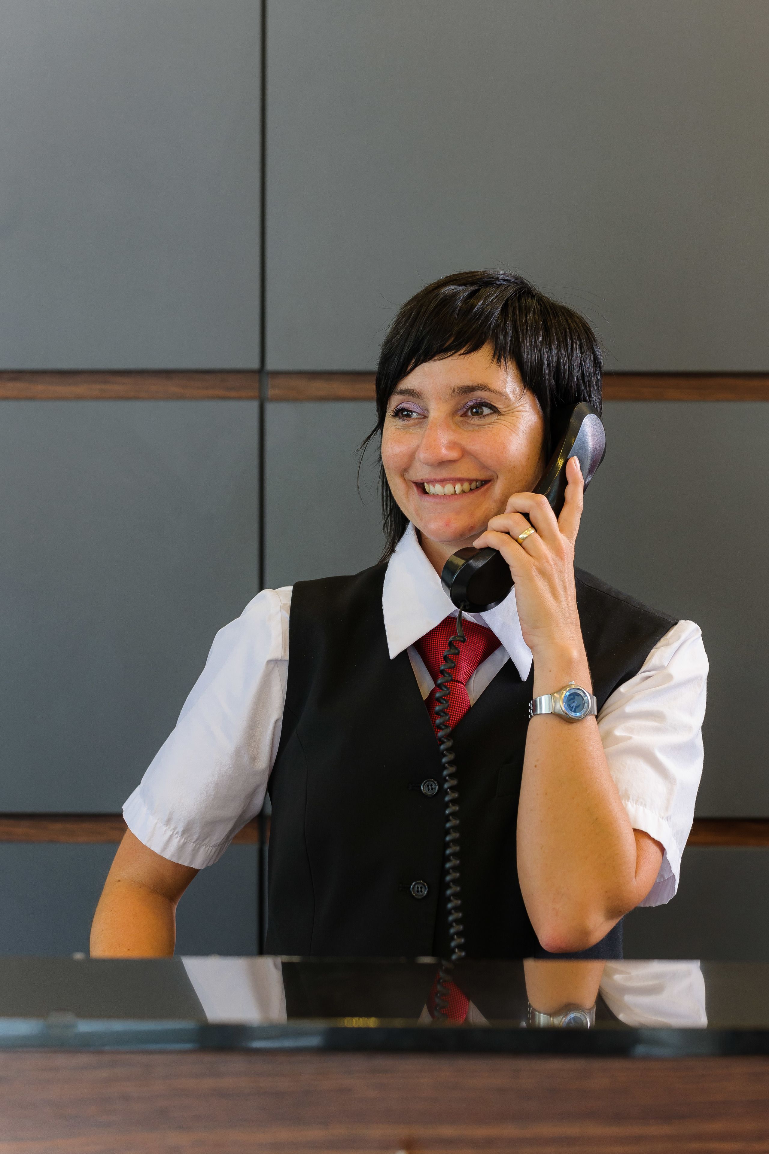 A person in a white shirt, black vest, and red tie sitting at a desk with a telephone.