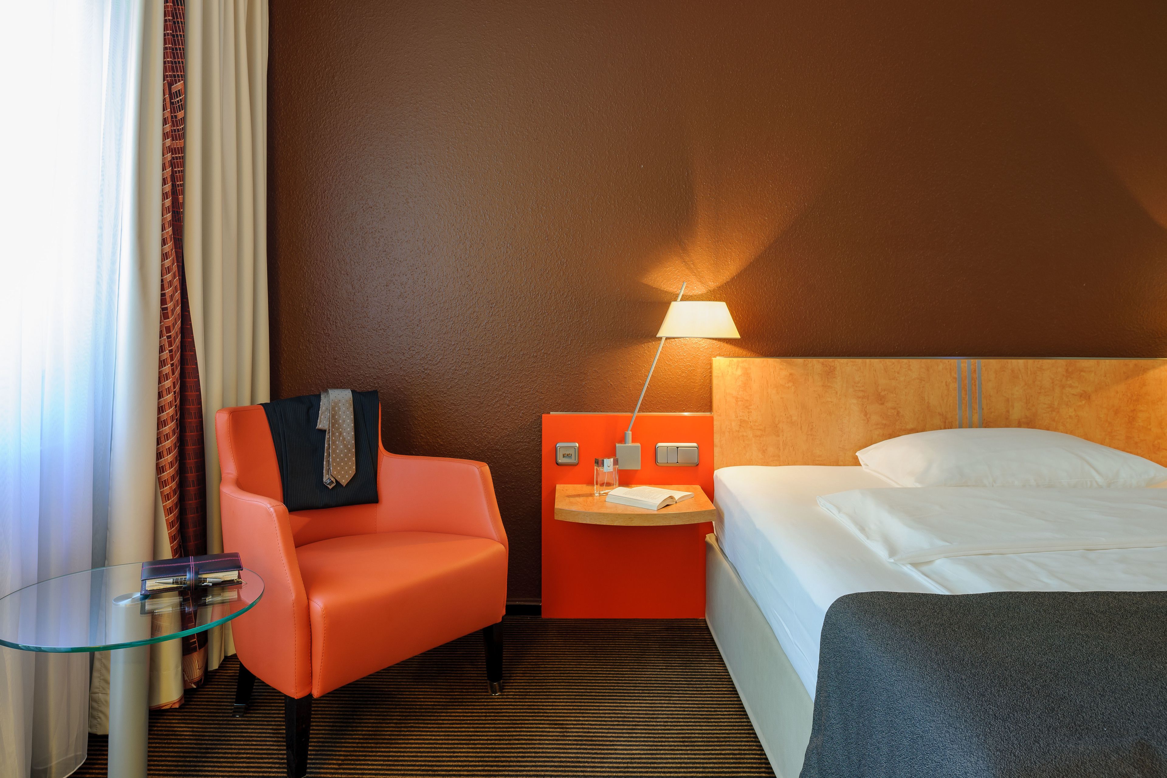 A hotel room with a bed, a glass table, an orange chair, and a nightstand with a lamp.